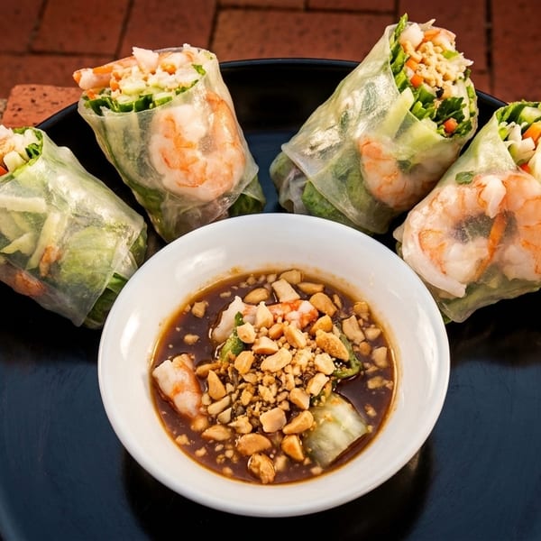 Four shrimp spring rolls with vegetables and a bowl of peanut dipping sauce on a black plate.