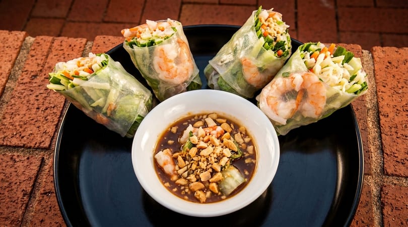 Four shrimp spring rolls with vegetables and a bowl of peanut dipping sauce on a black plate.